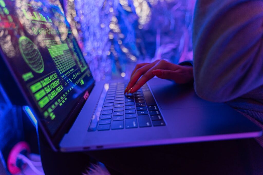 pexels-photo-5475752 Close-up of hands typing on a laptop displaying cybersecurity graphics, illuminated by purple light.